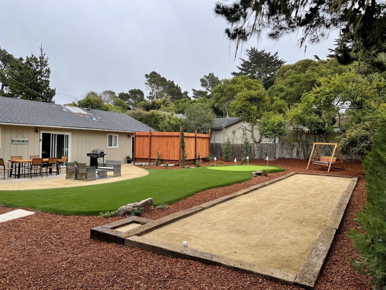 A backyard with a lawn, a sandpit, a wooden swing set, and a barbecue area.