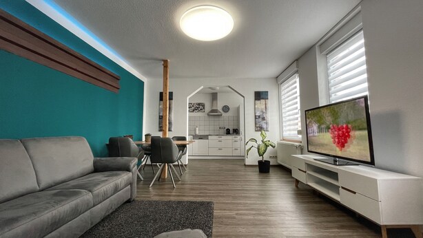 Una sala de estar moderna con un sofá gris, un comedor con sillas, una cocina al fondo y un televisor a la derecha.