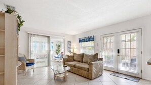 A spacious living room with a beige sofa, a glass coffee table, and a wicker chair. There are large windows with blinds, a door with a mat, and a staircase leading to another room. - Apt#1-Charming relaxing -beautiful APT-80yard walk to the BEACH wBBQ/ Patio (Pompano Beach)
