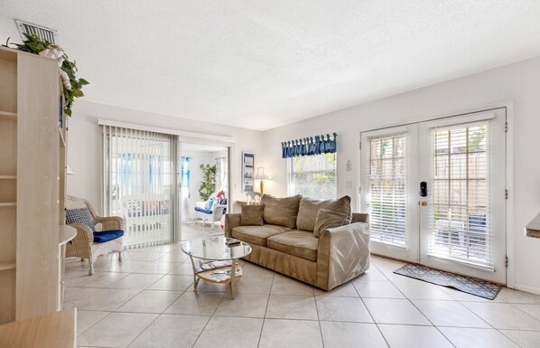 A spacious living room with a beige sofa, a glass coffee table, and a wicker chair. There are large windows with blinds, a door with a mat, and a staircase leading to another room.