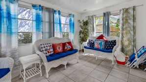A sunlit room with two white wicker sofas, one with red and white pillows, and another with blue cushions. There are blue curtains on the windows, and a ceiling fan is visible. - Apt#1-Charming relaxing -beautiful APT-80yard walk to the BEACH wBBQ/ Patio (Pompano Beach)