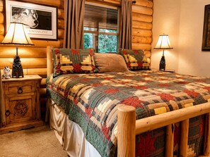 A log cabin-style bedroom with a wooden bed, two bedside tables with lamps, a framed picture on the wall, and a window with curtains. - Secluded Mountain Lodge with Game Room (Cloudcroft)