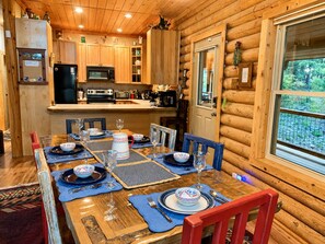Interior de una cabaña de madera con una mesa de comedor para seis comensales, una cocina con electrodomésticos modernos y un ventanal con vistas a follaje.