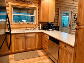 Cocina de cabaña de madera con armarios de madera, lavavajillas, cafetera y ventana con vista a la vegetación.
