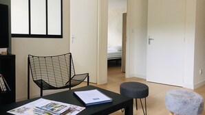 A room with a black chair, a small round stool, a table with a folder and papers, and a bed in the background.