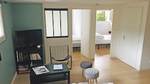 A living room with a sofa, a small table with papers, a bookshelf, a chair, and a view of a bedroom through an open door.