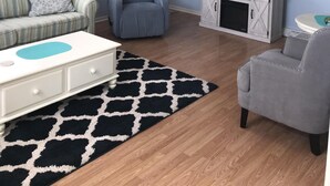 Een woonkamer met een plafondventilator, een televisie op een witte kast, een grijze fauteuil, een gestreepte bank en een zwart-wit kleed.