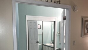 Een kamer met een deur naar een badkamer, een bed en een bord aan de muur met de tekst 'sun sand surf'.