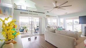 A spacious living room with a high ceiling fan, a sofa, and a view of the city through large windows. - Stunning Harbor Views! Boat Slip, 2 King Bedrooms, (Carolina Beach)
