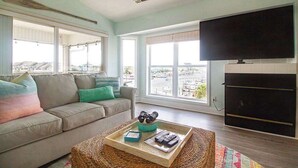 A living room with a sofa, a coffee table with remote controls, and a TV mounted on the wall. - Stunning Harbor Views! Boat Slip, 2 King Bedrooms, (Carolina Beach)