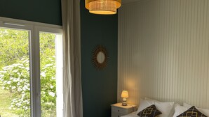 A bedroom with a bed, a nightstand, a lamp, a wall mirror, and a view of greenery through a sliding door. - Pretty house in a large garden / town center,  (La Flotte)