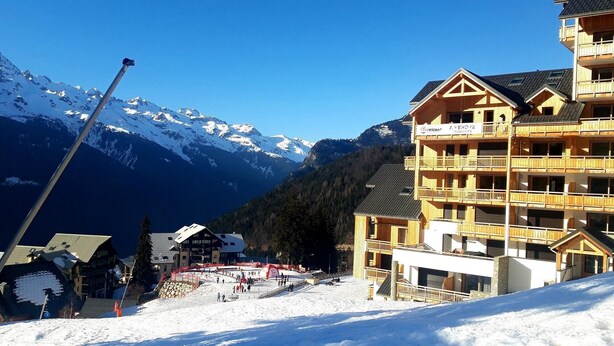 A multi-story wooden building with balconies, a ski slope in the foreground, and snow-covered mountains in the background.
