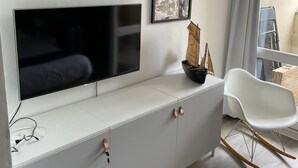 Uma sala de estar moderna com uma televisão de tela plana presa a um móvel branco, uma poltrona de balanço e uma pequena mesa com um modelo de navio.