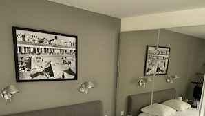 Uma cama bem arrumada com lençóis brancos e um cobertor azul-escuro, uma mesa de cabeceira com abajur e uma fotografia em preto e branco emoldurada na parede.