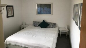 Ein Schlafzimmer mit einem Bett, zwei Nachttischen, einem Schranken und einem Bild an der Wand, sowie einem Fenster mit Blick auf Bäume.