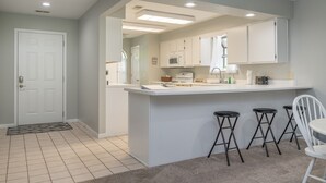 Una cucina moderna con mobili bianchi, un'isola centrale con sgabelli e un'apertura che dà su un'altra stanza.