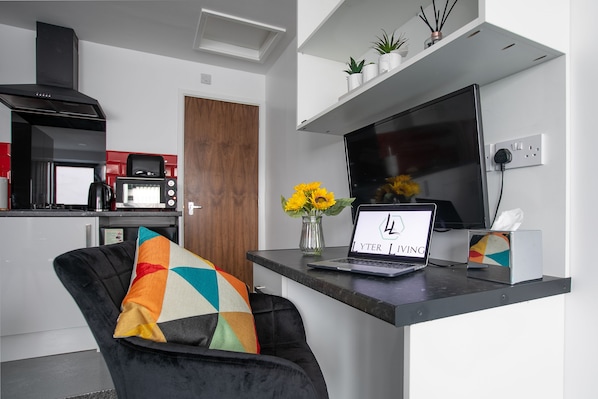 Une cuisine moderne équipée d’un four à micro-ondes, d’une machine à café et d’une hotte noire. On peut voir un ordinateur portable sur un bureau, ainsi qu’un vase rempli de fleurs jaunes et une boîte de mouchoirs.