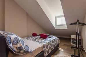 Een slaapkamer met een schuine kap, een bed met beddengoed met een patroon, een klein raam en een opslagkar.