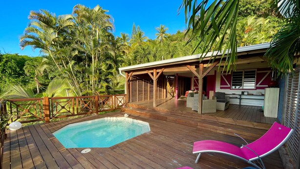 A wooden deck with a pool, lounge chairs, and a covered seating area with a view of tropical trees.