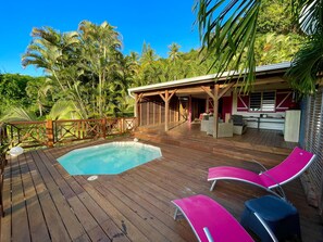 A wooden deck with a pool, lounge chairs, and a covered seating area with a view of tropical trees.