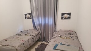 A small, neatly arranged bedroom with two single beds, a grey furry rug, and two wall-mounted pictures of white flowers. - Apparemment Agréable Idéal Pour Famille (Agadir)