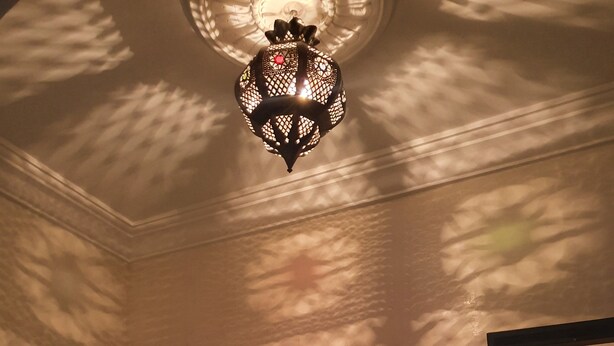 A hallway with a decorative ceiling light, a mirror, and a view into a kitchen area with a coffee machine and a patterned rug. - Apparemment Agréable Idéal Pour Famille (Agadir)