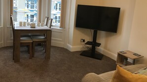 Una sala de estar con televisor, una mesa de comedor con sillas y una ventana con vista a edificios.