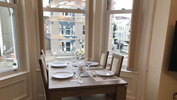 Una mesa de comedor para cuatro personas, con sillas, ubicada en un rincón de una habitación con ventanas amplias.