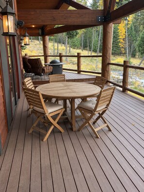 Una terraza de madera con una mesa redonda y sillas, rodeada por barandilla de madera, con vistas a un área boscosa.