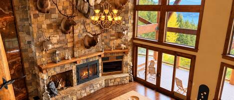 A cozy living room with a stone fireplace, mounted animal heads, and large windows offering a view of the outdoors.