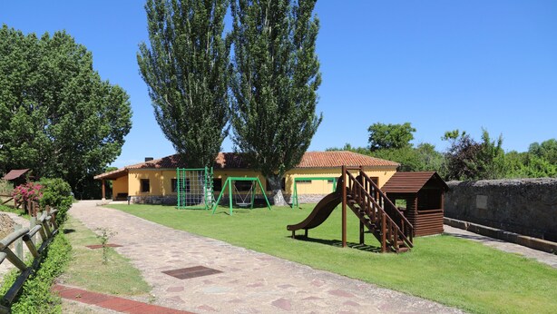 Ein Spielplatz mit Rutsche und Schaukeln, ein Pflasterweg und ein Gebäude mit Ziegeldach.