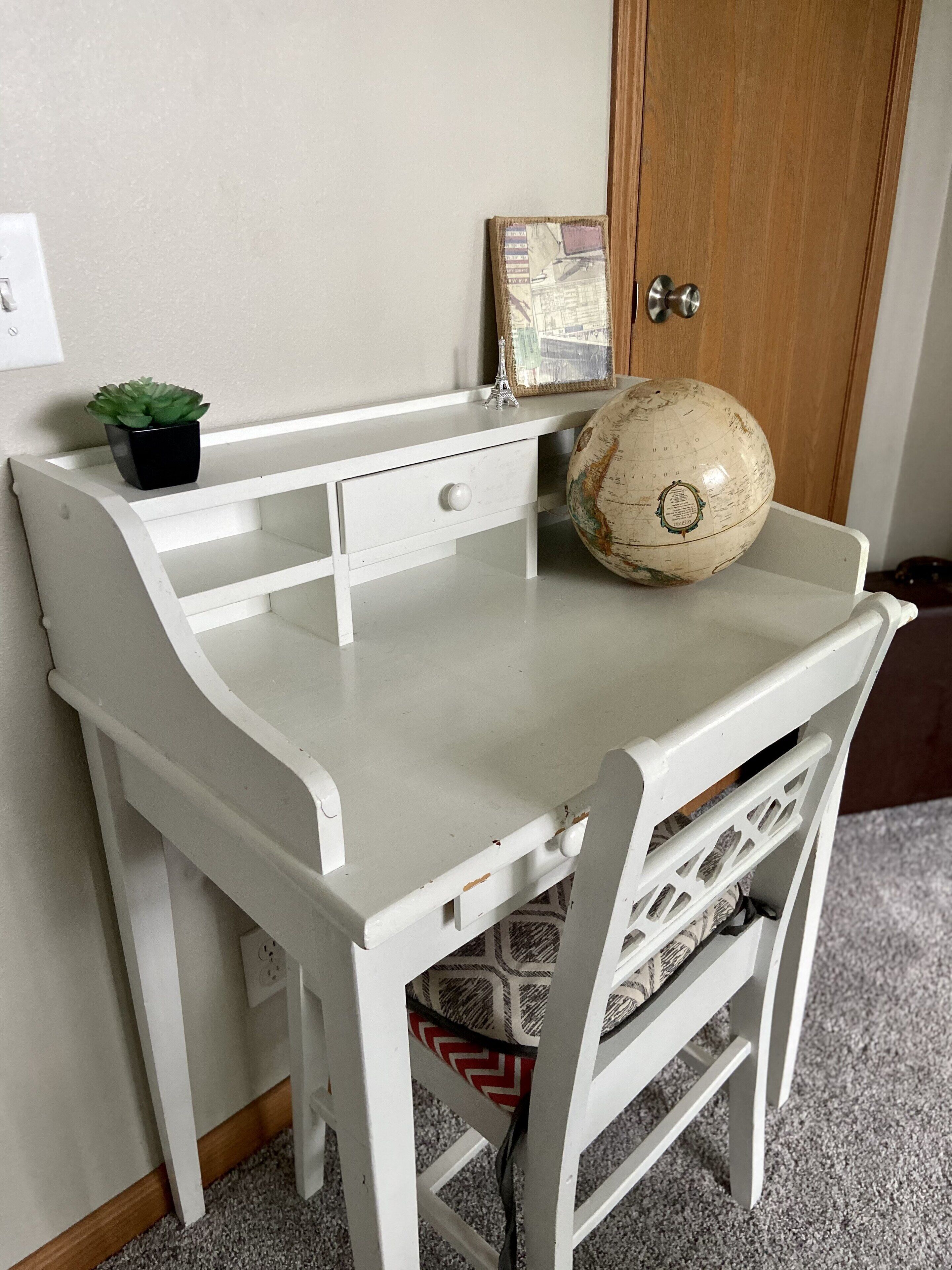 Una scrivania in legno bianco con un mappamondo, una piccola pianta in vaso e una cornice con un'immagine.
