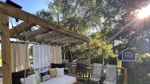 Una terrassa de fusta amb una cadira hamaca, una barbacoa i un cobert petit.