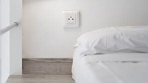 Un dormitorio con pared blanca, un enchufe, suelo de madera y una cama con ropa blanca.