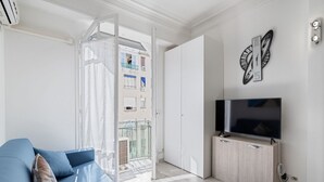 Una sala de estar moderna con un sofá azul, un televisor sobre un mueble de madera y un ventanal con vistas a un balcón y edificios.