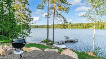 A lakeside camping area with a barbecue grill, a dock with a boat, and a bench.