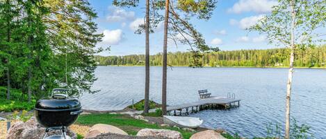 Un área de camping junto al lago, con una parrilla, un muelle con un bote y una banca.