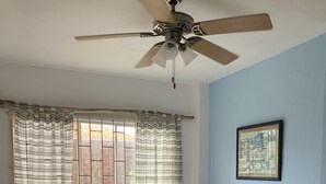 Une chambre à coucher avec un lit, un ventilateur de plafond, une fenêtre avec des rideaux et un tableau encadré au mur.