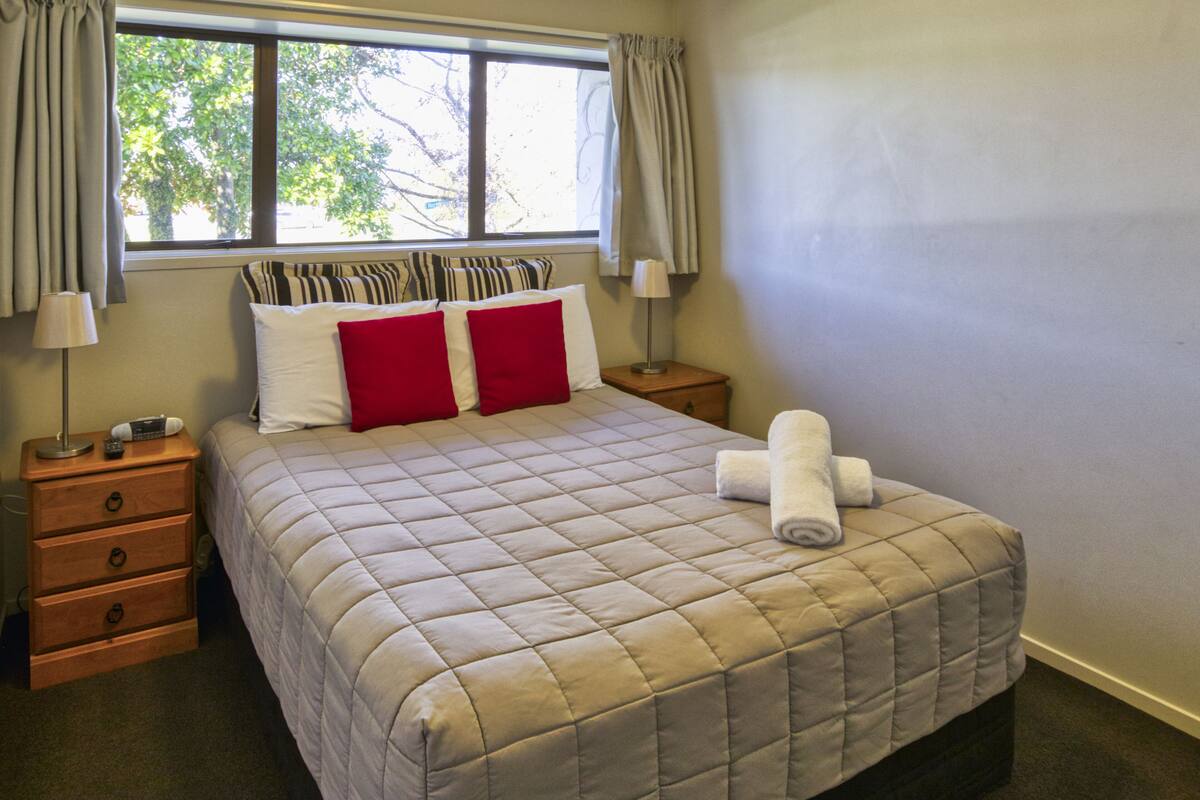 Ein ordentlich bezogenes Bett mit einer gesteppten Decke, zwei roten Kissen und einem aufgerollten weißen Handtuch am Fußteil. Zwei Nachttische mit Lampen und Telefon sind vorhanden. Das Zimmer hat ein Fenster mit Vorhängen und Blick auf Bäume.