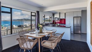 Une cuisine moderne avec des meubles blancs et un îlot central rouge, un coin repas avec des chaises en osier et une table dressée pour quatre personnes, ainsi qu’une grande fenêtre donnant sur une vue imprenable sur la côte.