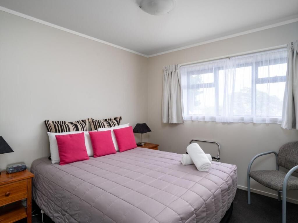 A bedroom with a bed, pillows, a chair, a nightstand, a lamp, and a window with curtains.