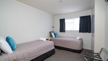 Habitación con dos camas, cada una con una almohada y una toalla enrollada. Se ve una ventana con cortinas, una mesita y una silla.
