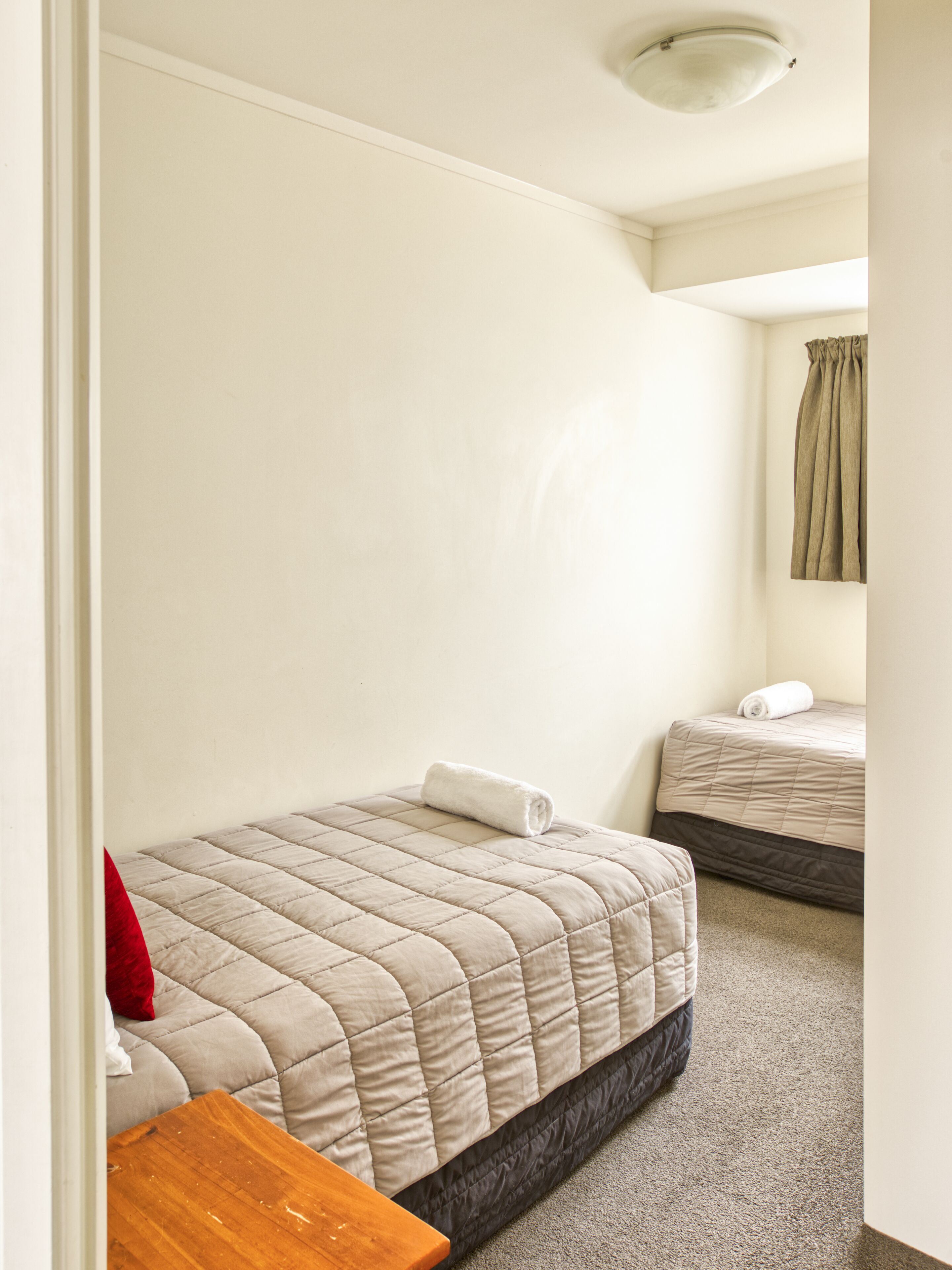 A bedroom with two beds, a wooden nightstand, a red pillow, and a ceiling light.