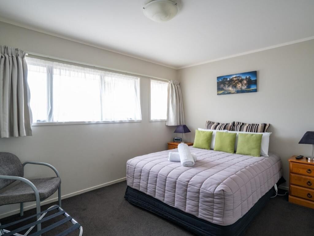 A bedroom with a bed, a chair, a nightstand, a lamp, a painting, and a window with curtains.