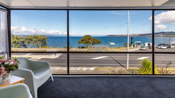 Une pièce dotée de grandes fenêtres donnant sur la mer, un espace de stationnement et quelques chaises.