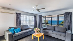 Un salon moderne avec deux canapés gris, une table basse en bois et un ventilateur de plafond. De grandes fenêtres offrent une vue sur la mer.