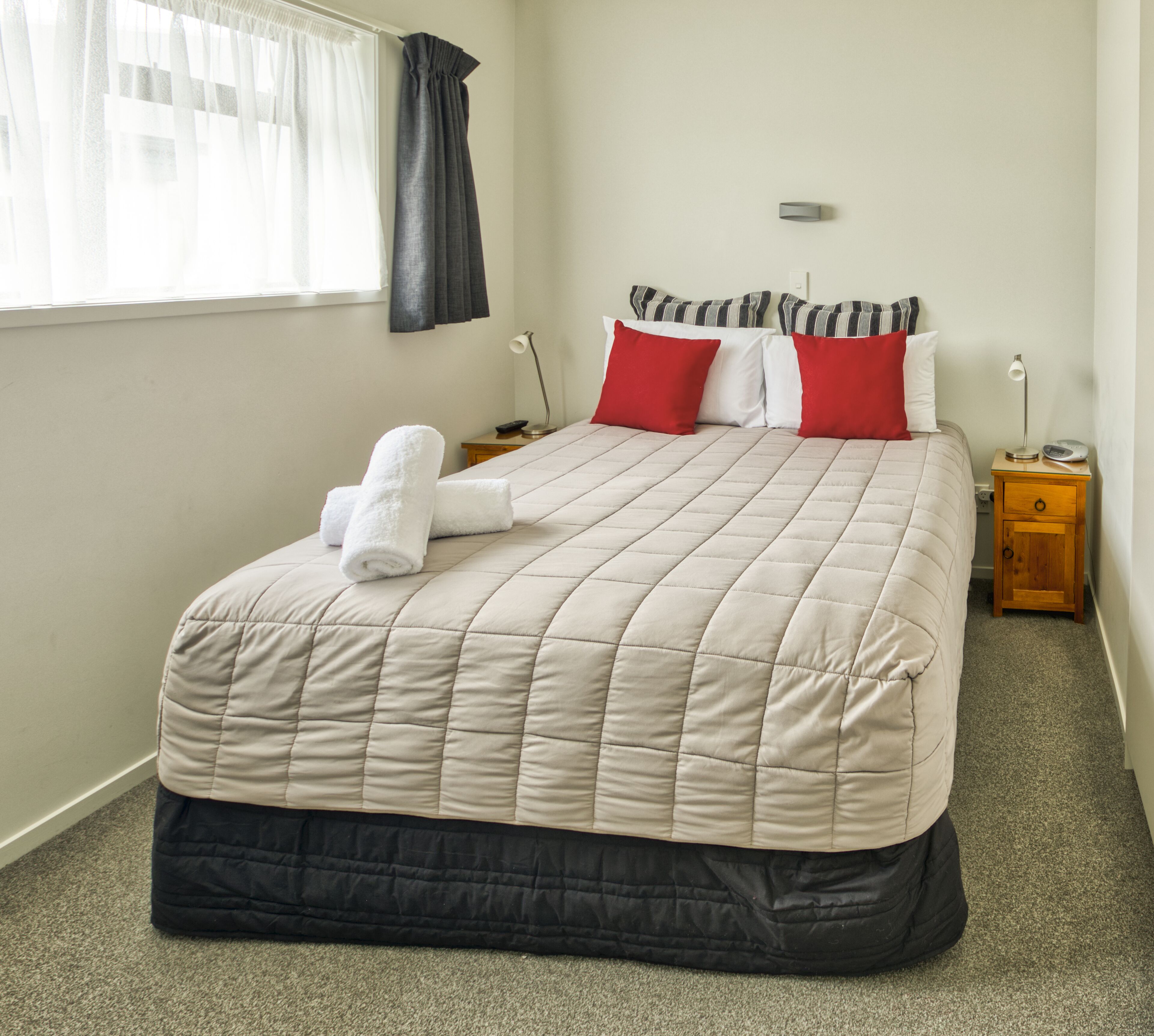 Cama bem arrumada com colcha bege, almofadas vermelhas e brancas e toalha branca. Há uma mesa de cabeceira com luminária e um pequeno relógio. O quarto tem uma janela com cortinas brancas e um ponto de luz fixado na parede.