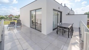 Un patio exterior moderno con una mesa y sillas de comedor, un ventanal de vidrio que da a un área cubierta y vistas del paisaje circundante.