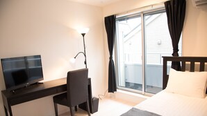 A hotel room with a bed, a desk with a TV, a chair, a lamp, and a window with curtains. - Basic Stay Sugamo (北区)