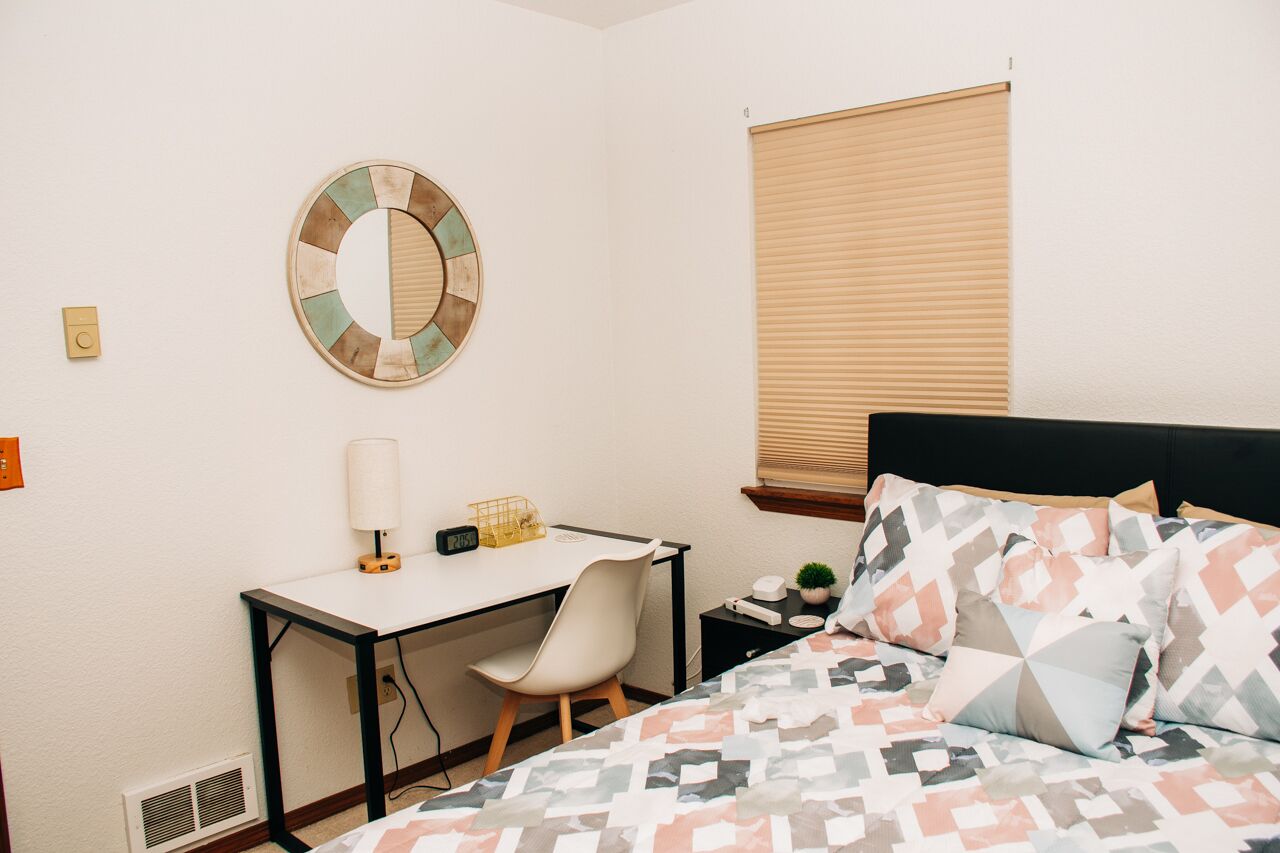 A bedroom with a bed, a desk, a chair, a lamp, a clock, a small plant, and a window with blinds.
